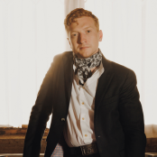 Man standing in front of window, wearing brown suit jacket and a neckerchief